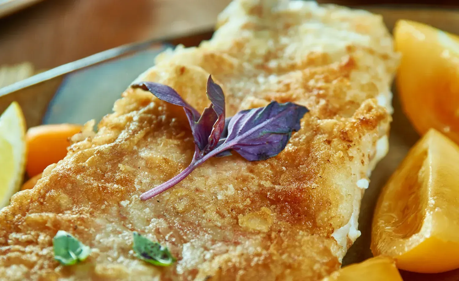 Filete de pescado frito crujiente en un plato con rodajas de tomate naranja y una ramita de albahaca morada.