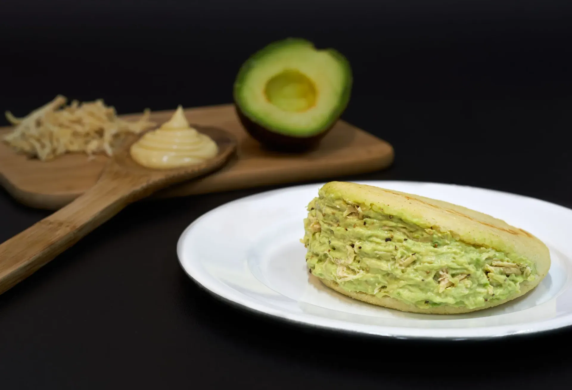 Arepa rellena de mezcla de aguacate verde sobre un plato blanco, con ingredientes en el fondo.