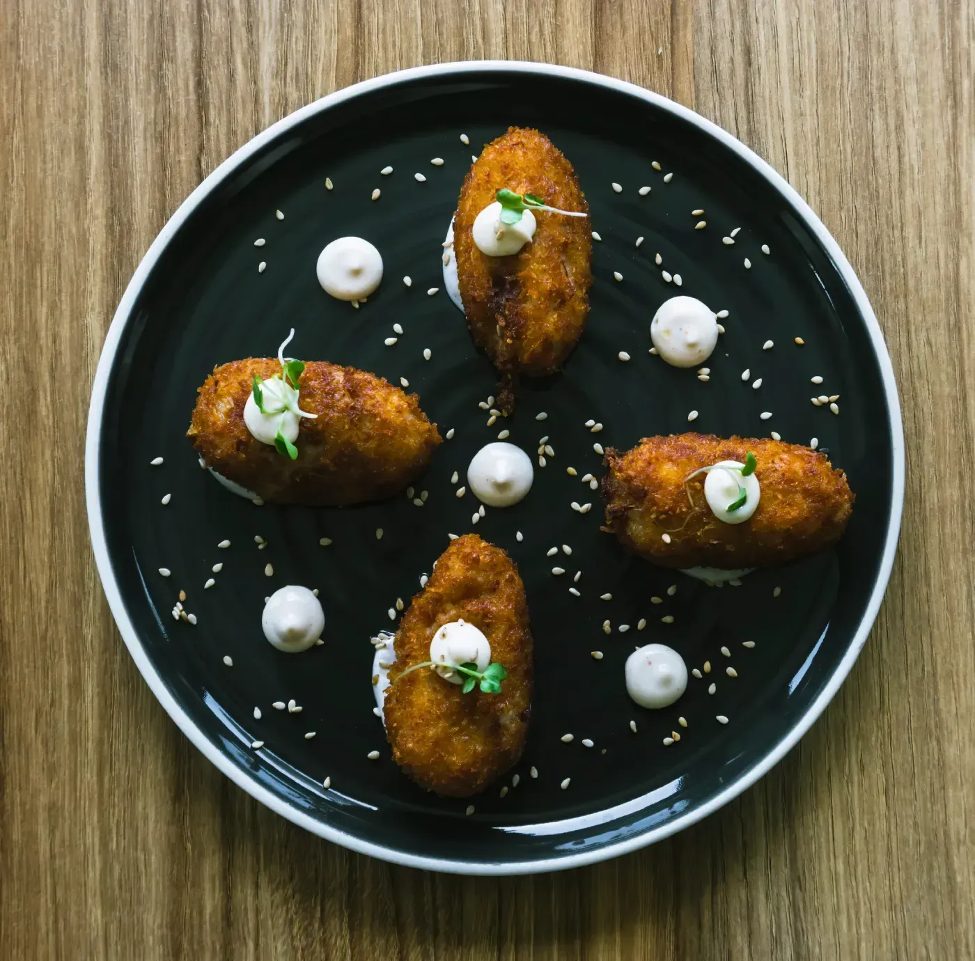 Cinco croquetas doradas con salsa blanca y semillas de sésamo en un plato negro, sobre una superficie de madera.