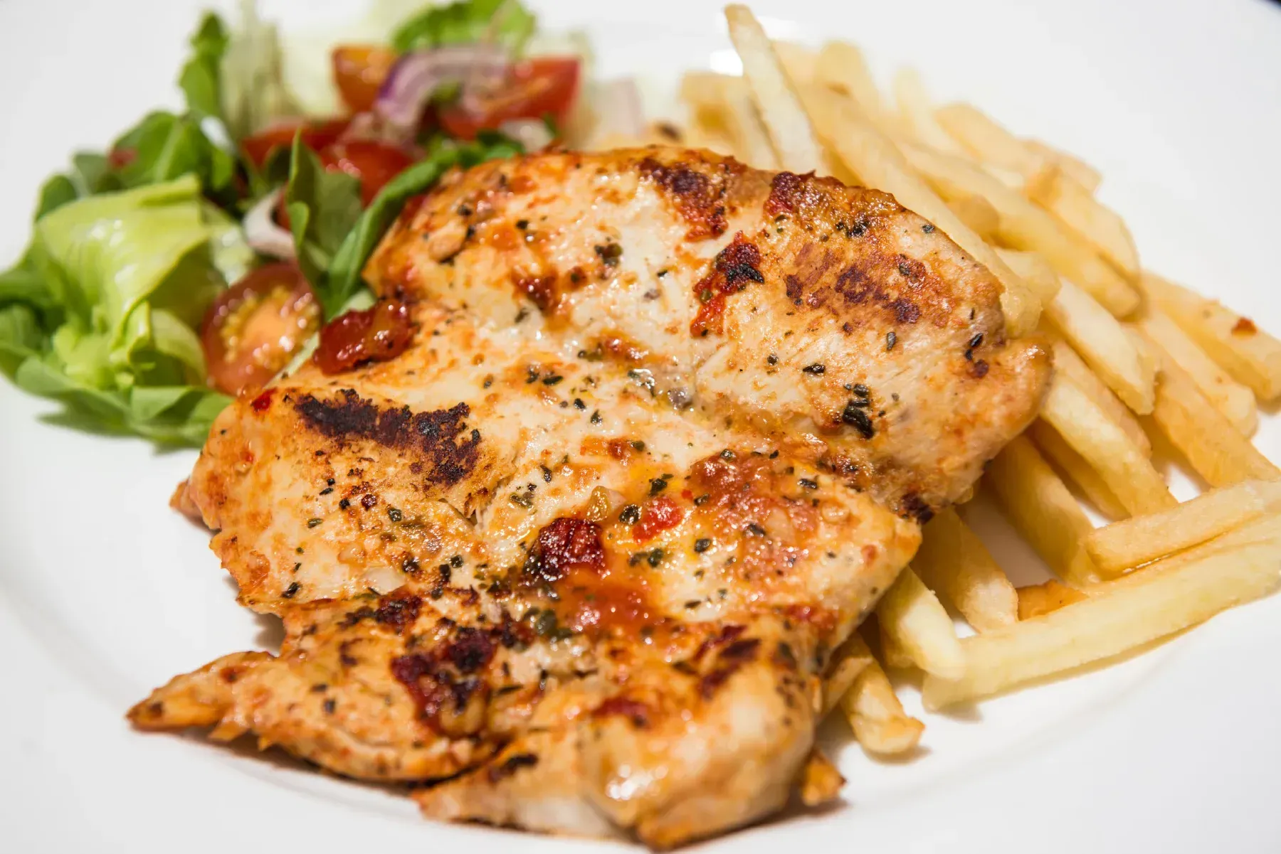 Pechuga de pollo a la parrilla con patatas fritas y ensalada en un plato blanco.