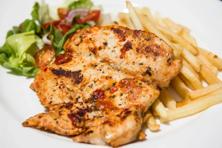 Pechuga de pollo a la parrilla con patatas fritas y ensalada en un plato blanco.