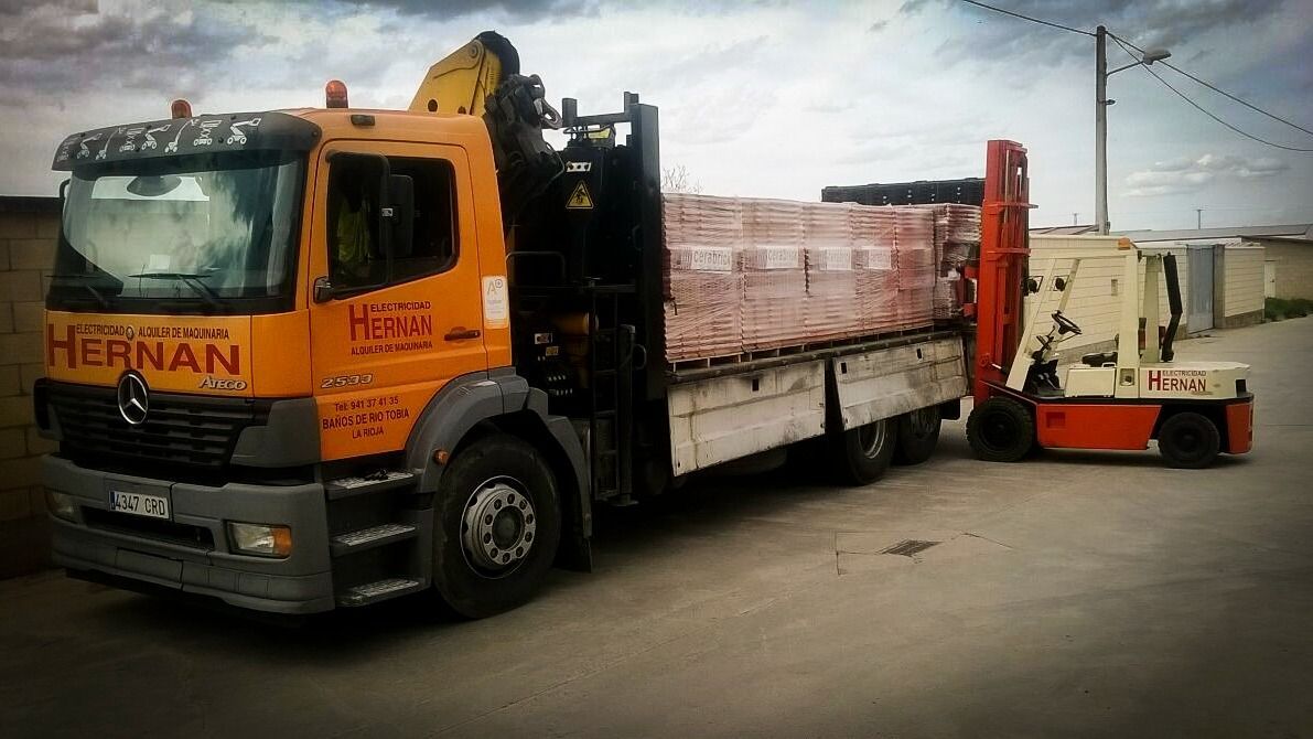 Un camión volquete naranja cargado de ladrillos remolca una pequeña carretilla elevadora roja en un patio industrial.
