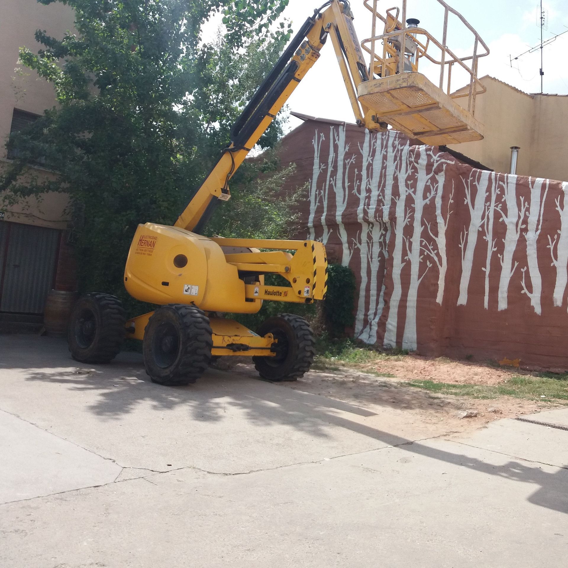 Plataforma elevadora amarilla junto a un muro de ladrillos y un árbol en una calle soleada.