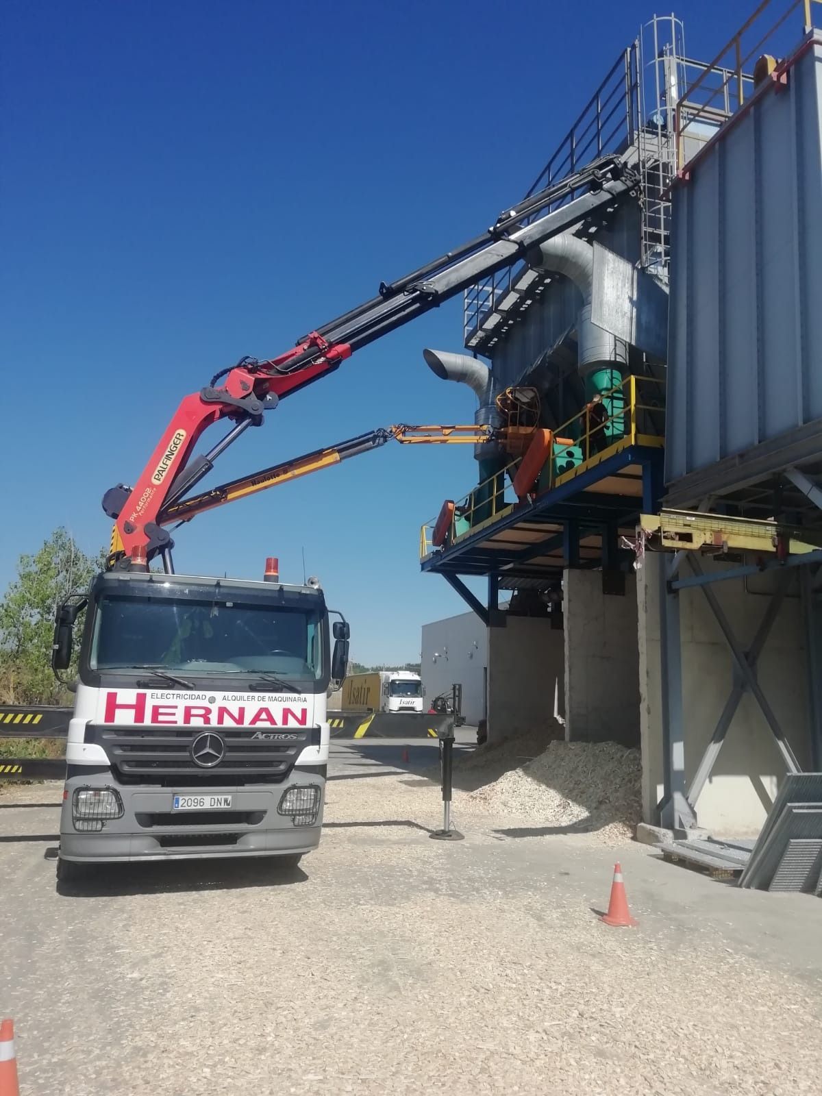 Un camión grúa levanta una estructura metálica junto a un edificio industrial elevado en un día soleado.