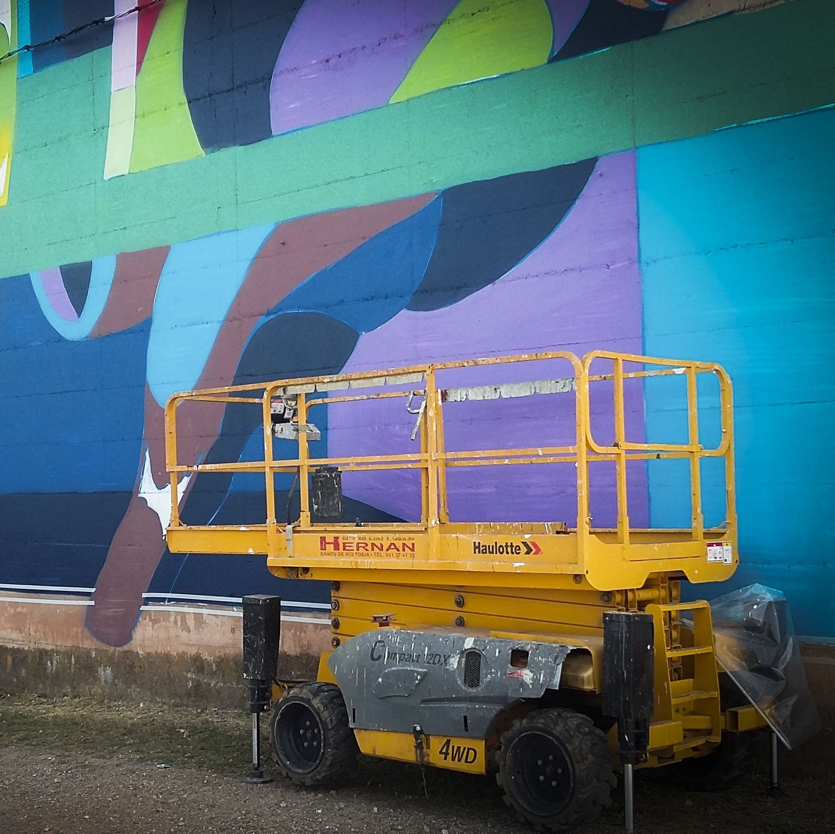 Una plataforma elevadora de tijera amarilla está estacionada junto a un mural colorido en el exterior.