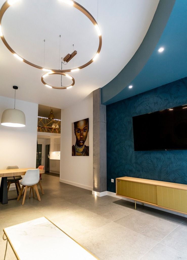 Sala de estar moderna con pared decorativa azul, televisor montado en la pared y área de comedor con lámparas colgantes y aros de luz.