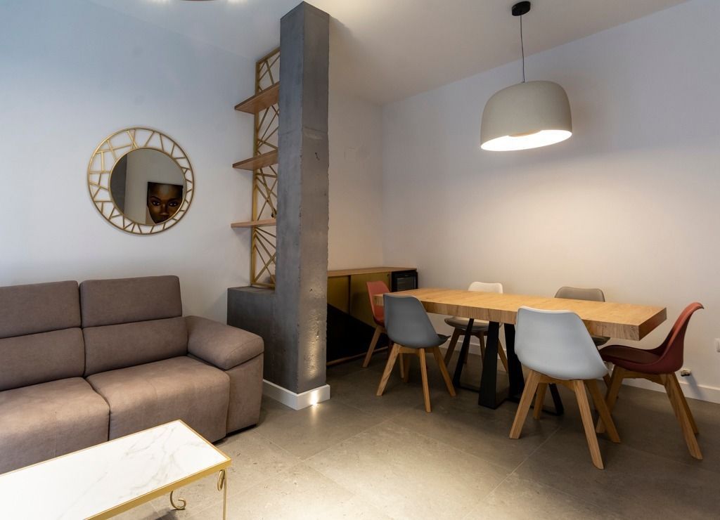 Salón-comedor moderno con sofá gris, espejo redondo, mesa de madera y sillas variadas bajo una lámpara colgante.