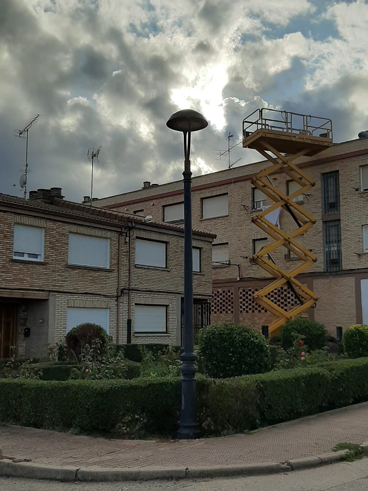 Edificio de ladrillo con una escalera de incendios amarilla bajo un cielo nublado, bordeado por setos cuidadosamente recortados.