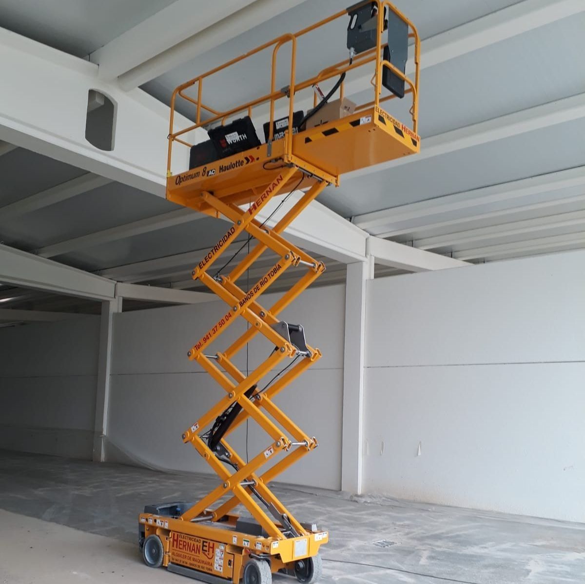 Una plataforma elevadora de tijera naranja se eleva bajo el techo de un almacén, alcanzando una viga de acero.