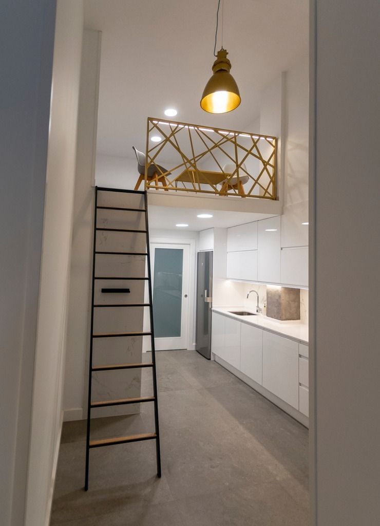 Cocina moderna estilo loft con escalera que da acceso a un altillo, armarios blancos y una lámpara colgante dorada.