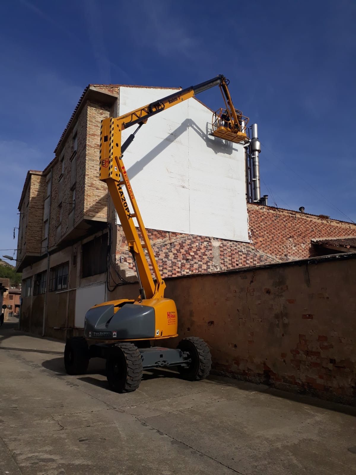 Plataforma elevadora amarilla que se eleva hasta la pared de un edificio en una calle estrecha bajo un cielo despejado.
