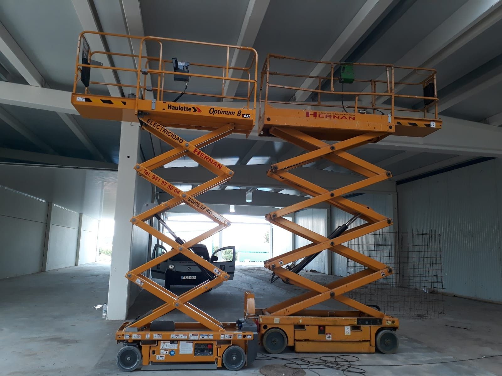 Una plataforma elevadora de tijera amarilla se eleva dentro de un almacén industrial vacío bajo un techo metálico.