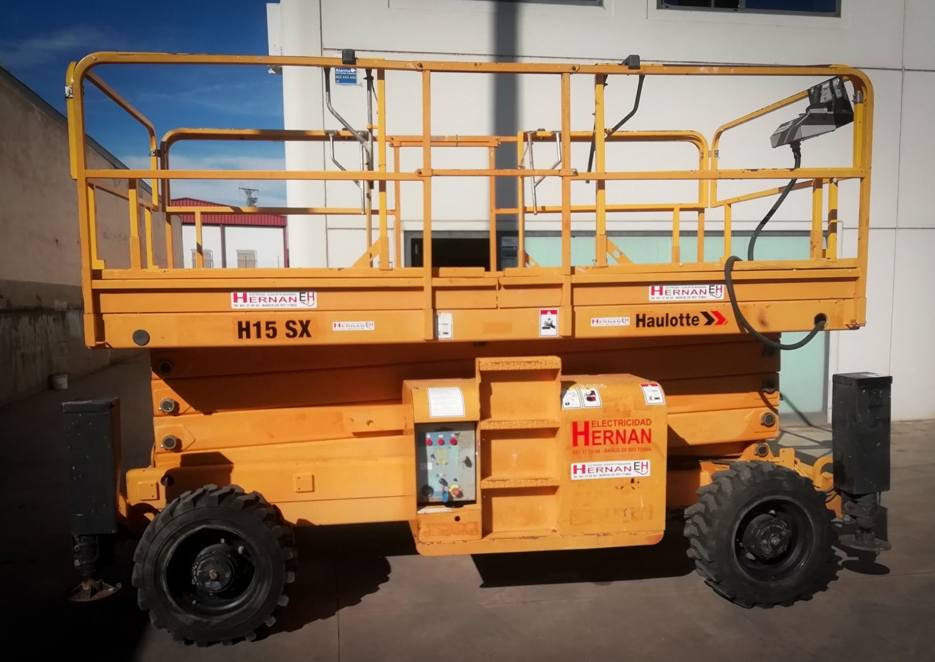 Plataforma elevadora de tijera Orange H15 SX estacionada al aire libre contra una pared.