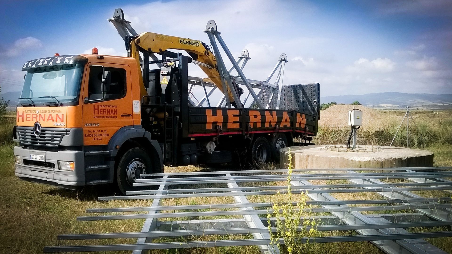 Camión naranja de la compañía HEBAN con grúa estacionado junto a estructuras metálicas apiladas en un campo rural.