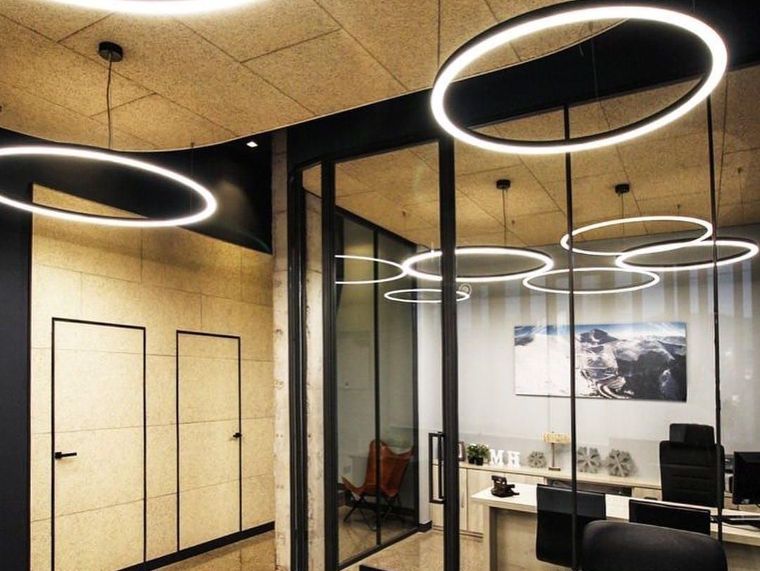 Sala de reuniones moderna con paredes de cristal, lámparas colgantes circulares y escritorios con sillas.