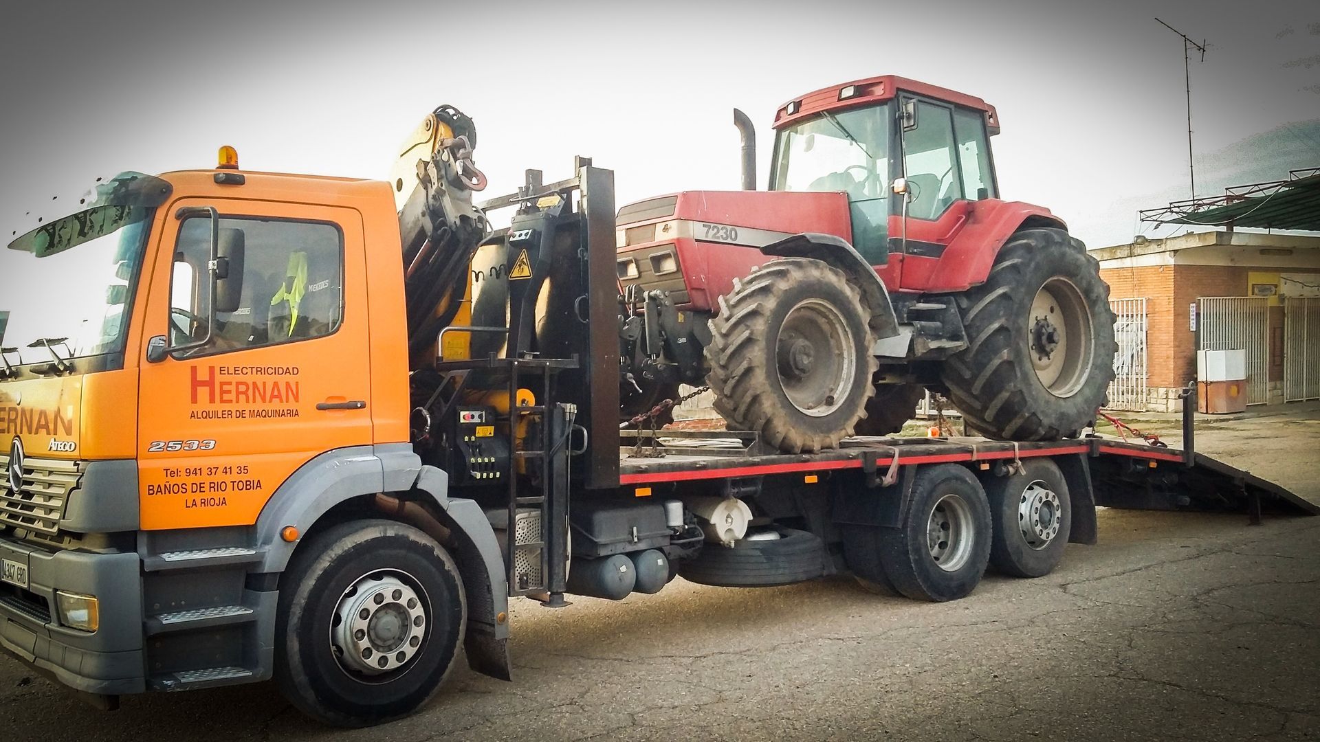 Grúa naranja remolcando un tractor rojo en un remolque de plataforma en un entorno de carretera.