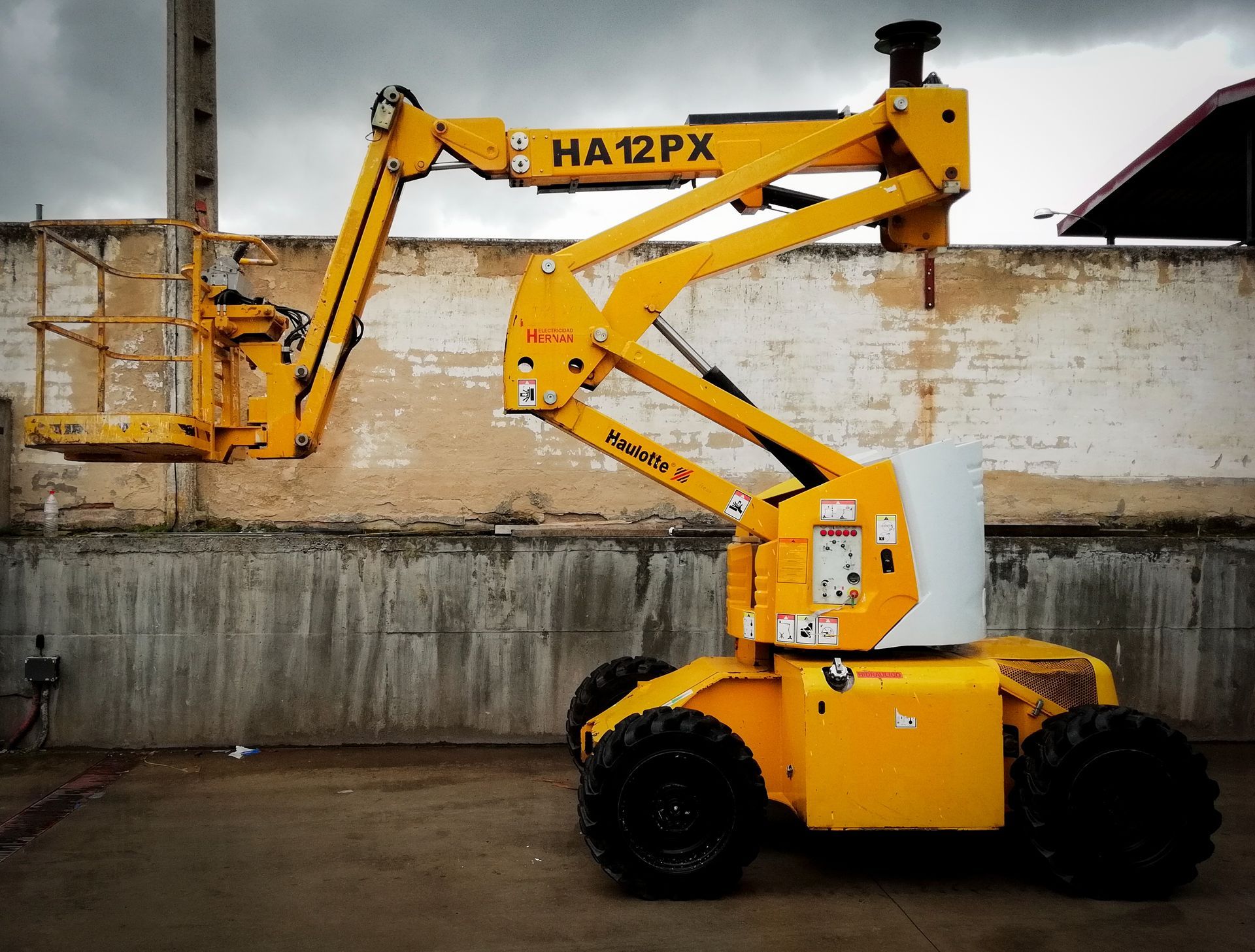 Una plataforma elevadora amarilla de construcción está estacionada junto a un muro de hormigón en un patio industrial.