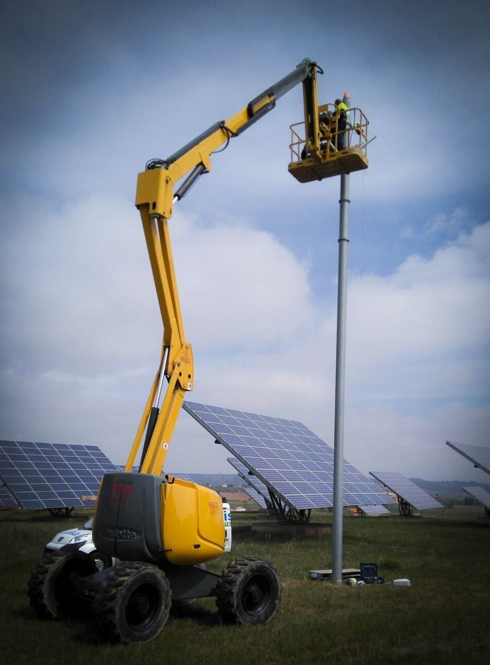 Una plataforma elevadora amarilla sube a los trabajadores junto a unos paneles solares en un campo de hierba.