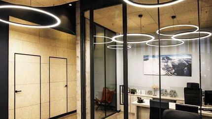 Interior de oficina moderno con paredes de cristal, luces de techo circulares, escritorios y sillas.