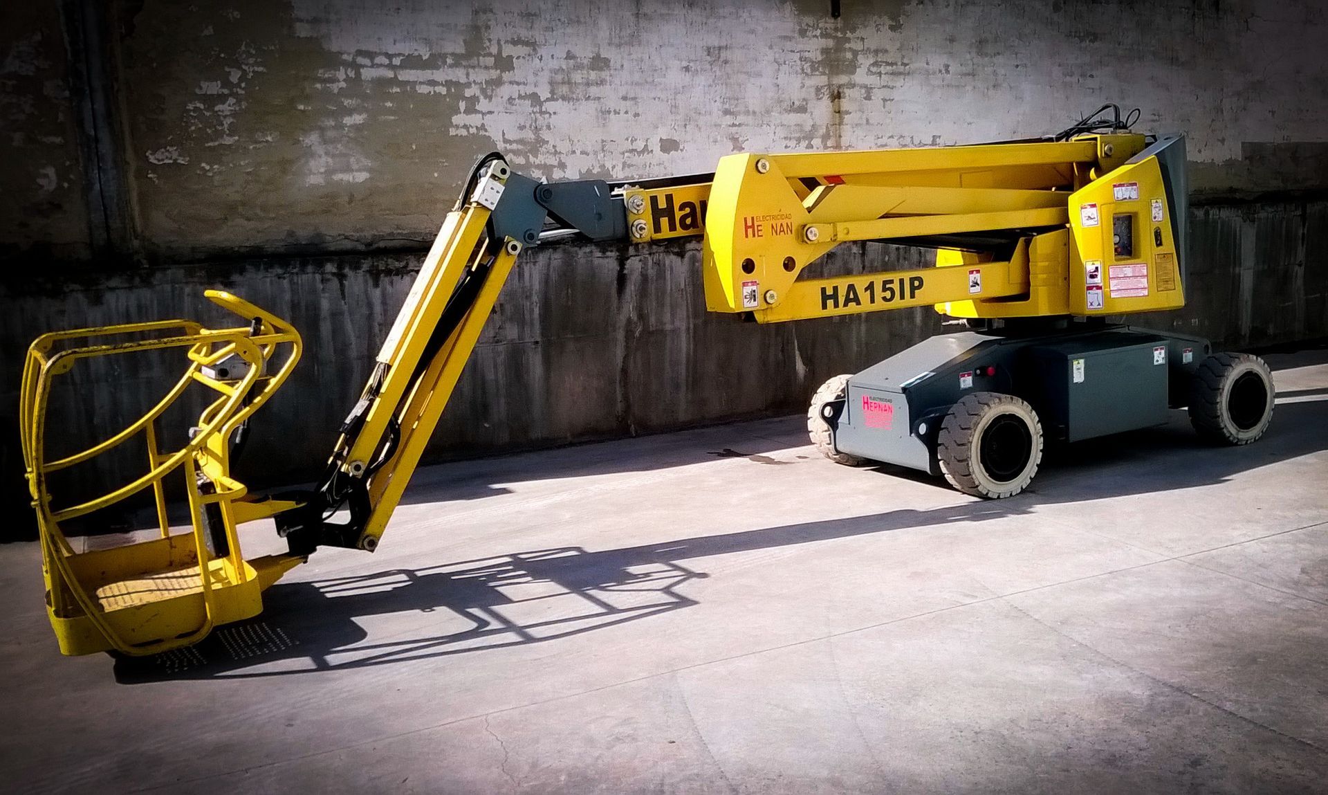 Una plataforma elevadora amarilla está estacionada en una zona industrial junto a un muro de hormigón.