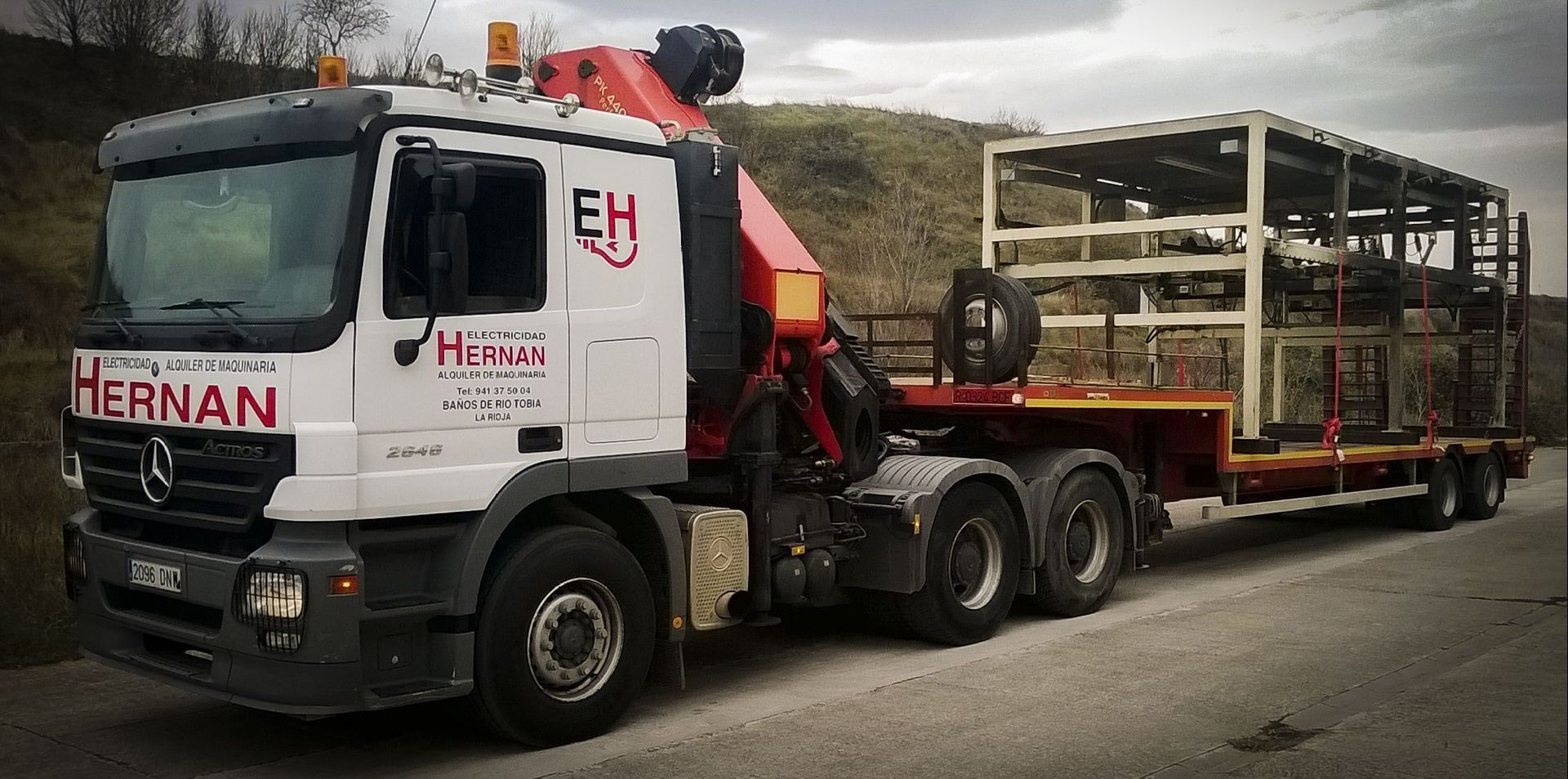 Camión blanco HERNAN remolcando un remolque de plataforma con estructura metálica al costado de la carretera, grúa montada detrás de la cabina.