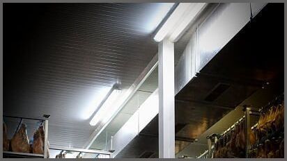 Luces fluorescentes en un techo corrugado en un pasillo de un mercado interior, con productos colgados a ambos lados.