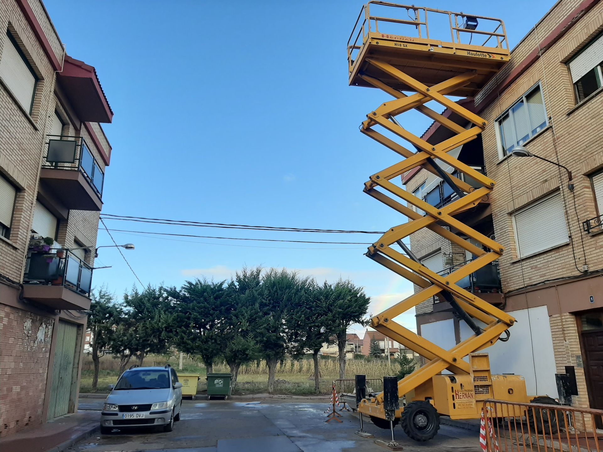 Una plataforma elevadora de tijera amarilla se eleva junto a un edificio de apartamentos en un patio interior, con una furgoneta y árboles cerca.