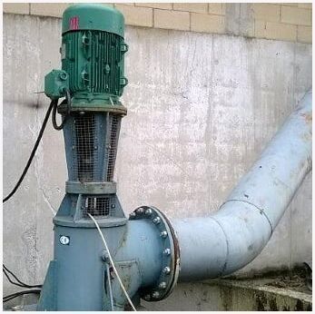 Bomba industrial con motor verde conectada a una gran tubería gris contra una pared de hormigón.