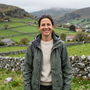 Persona sonriente con una chaqueta verde junto a un muro de piedra en un valle rural rodeado de colinas y campos.