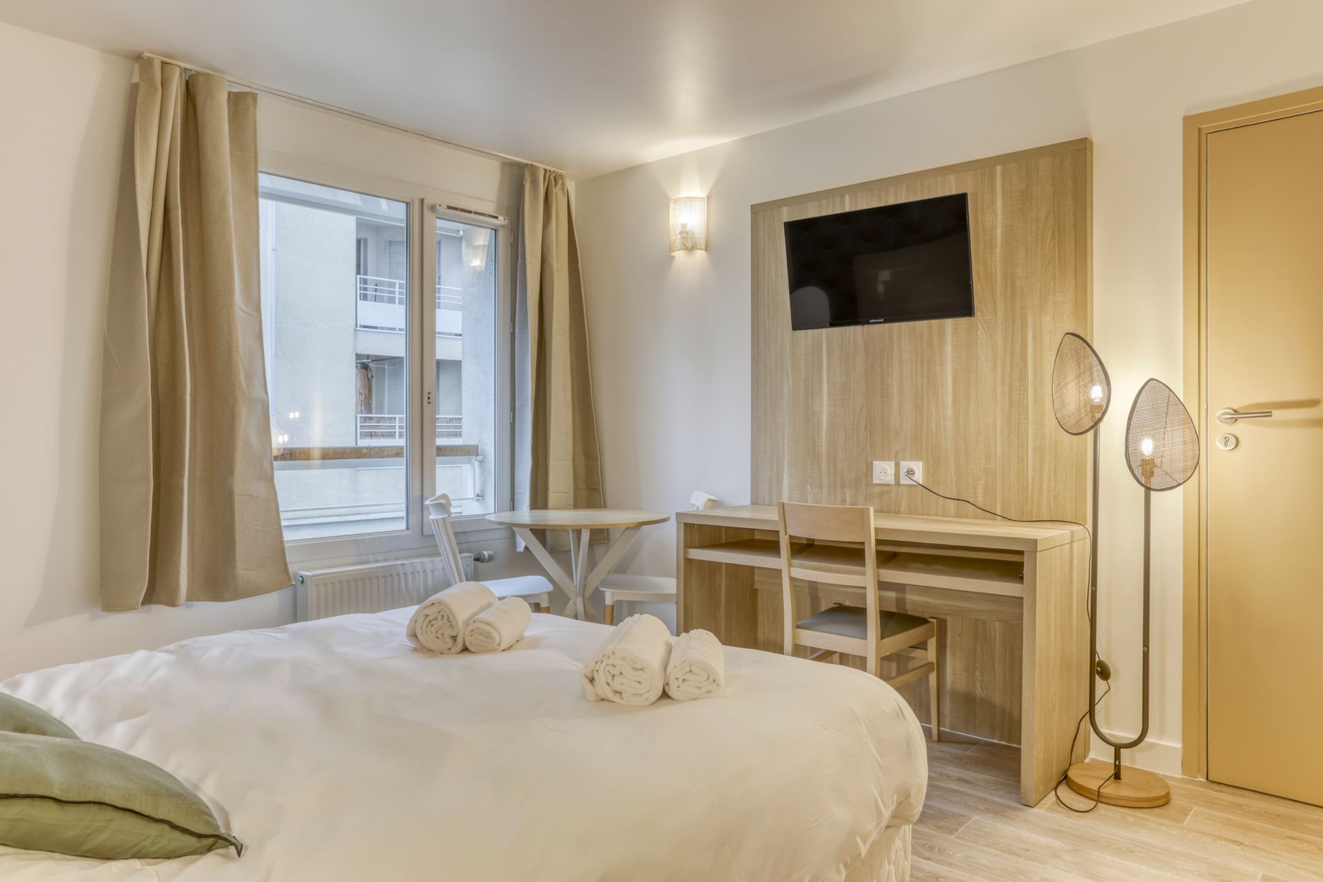 Résidence Colivi, Appart Hôtel à Annemasse. Chambre lumineuse avec lit, bureau, fenêtre et télévision murale dans un intérieur beige minimaliste