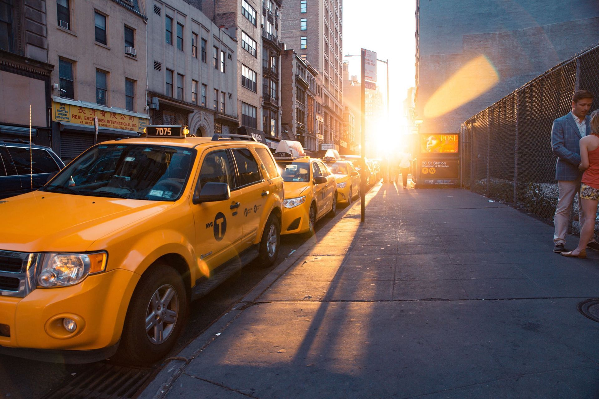 Taxis amarillos estacionados en una calle de la ciudad, con el sol poniéndose entre los edificios. Una pareja está de pie en la acera.
