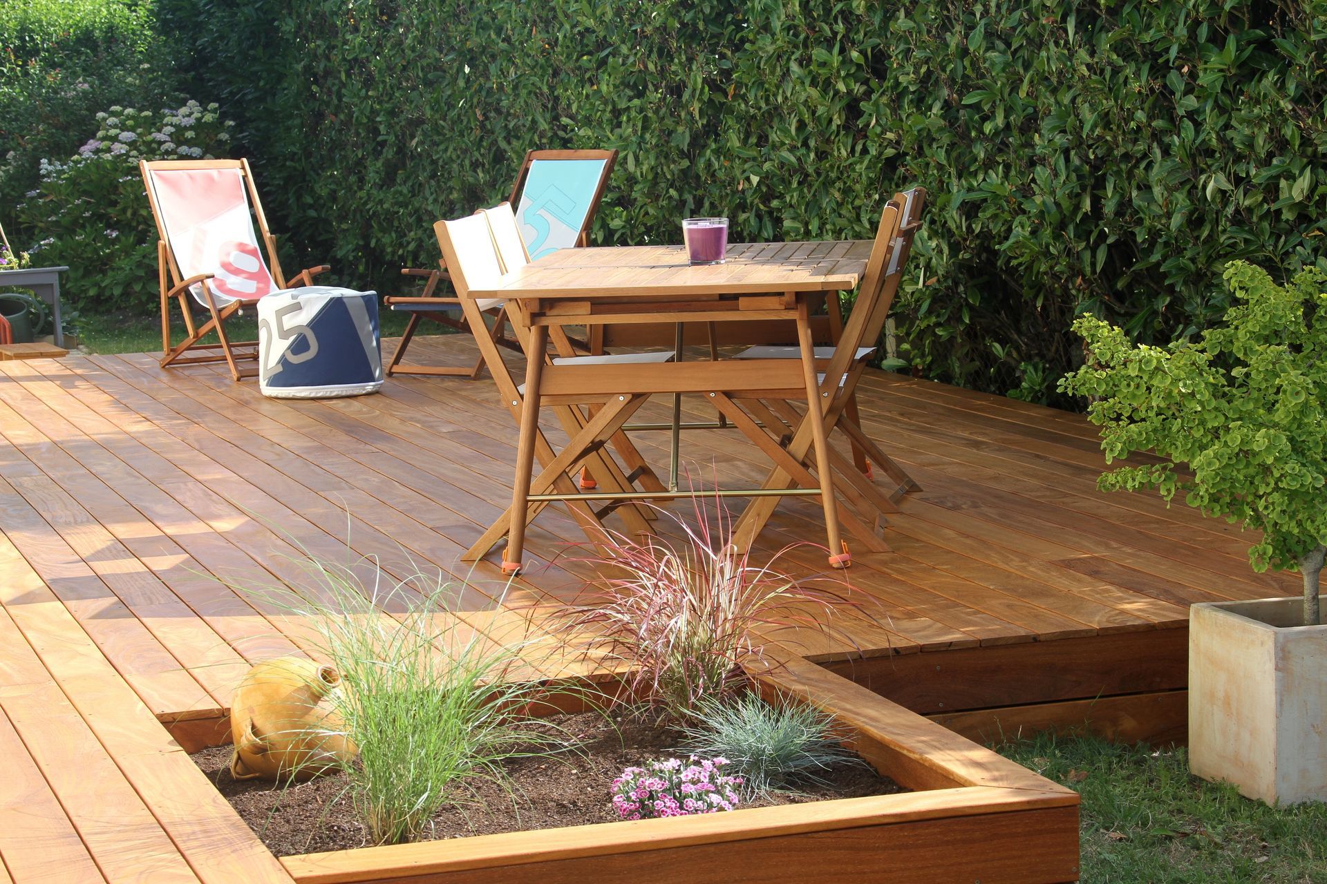 Une terrasse en bois avec une table, des chaises, un transat et une petite jardinière, adossée à une haute haie verte.