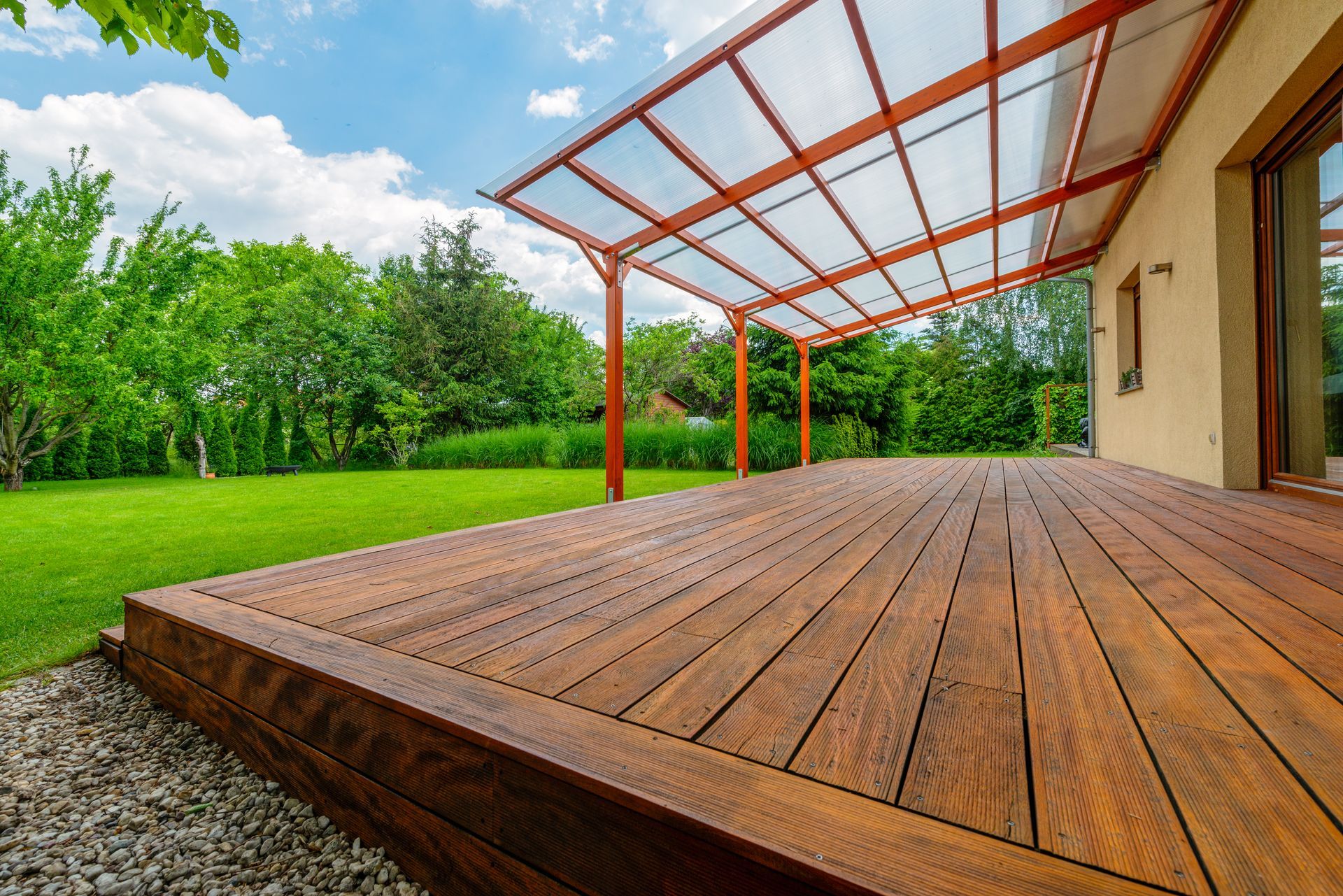 Une terrasse en bois avec une extension de toit translucide en pente, attenante à une maison et donnant sur une pelouse verdoyante.