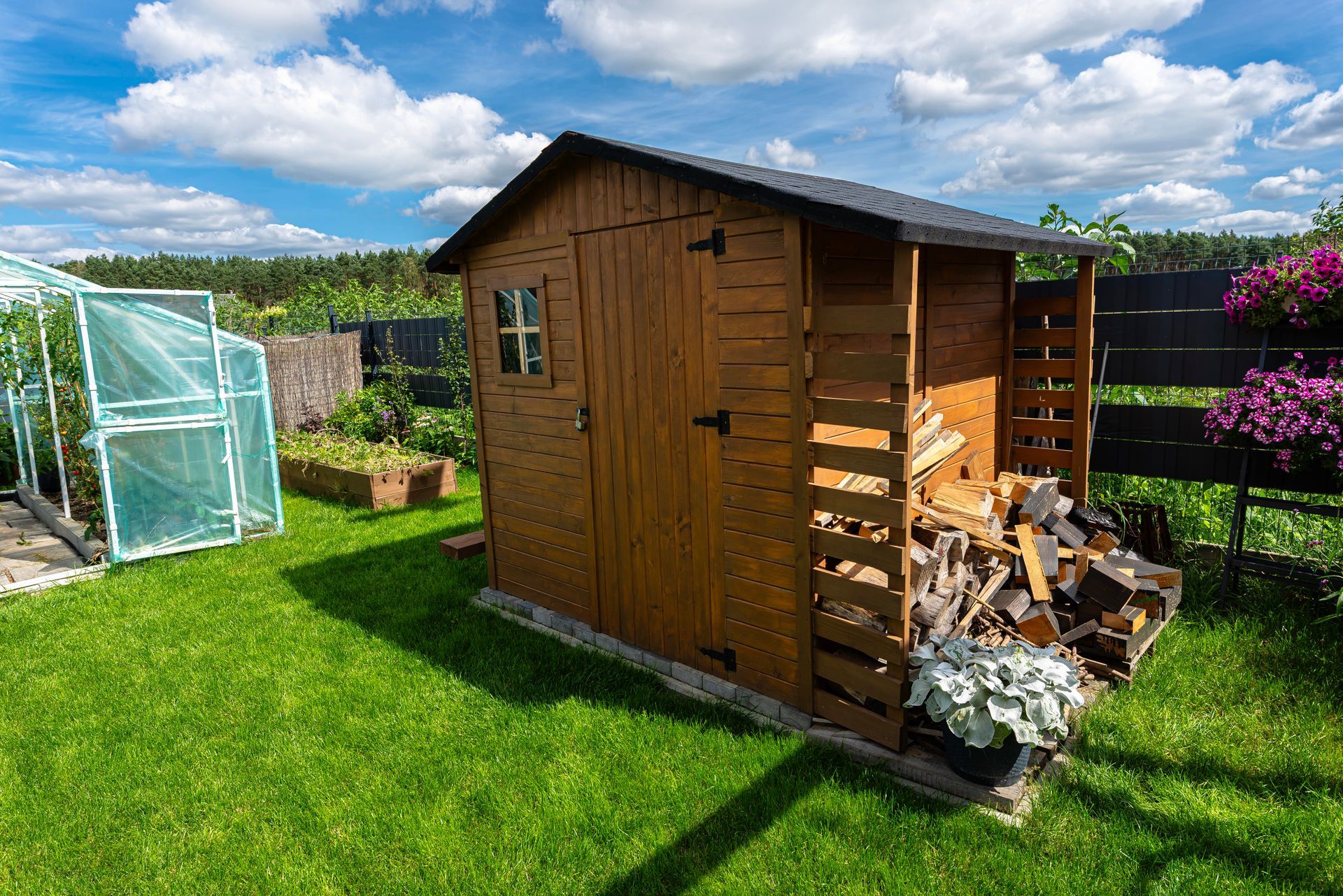 Un abri de jardin en bois, avec une pile de bûches attenante, se dresse sur une pelouse verte, à côté d'une petite serre, sous un ciel bleu.
