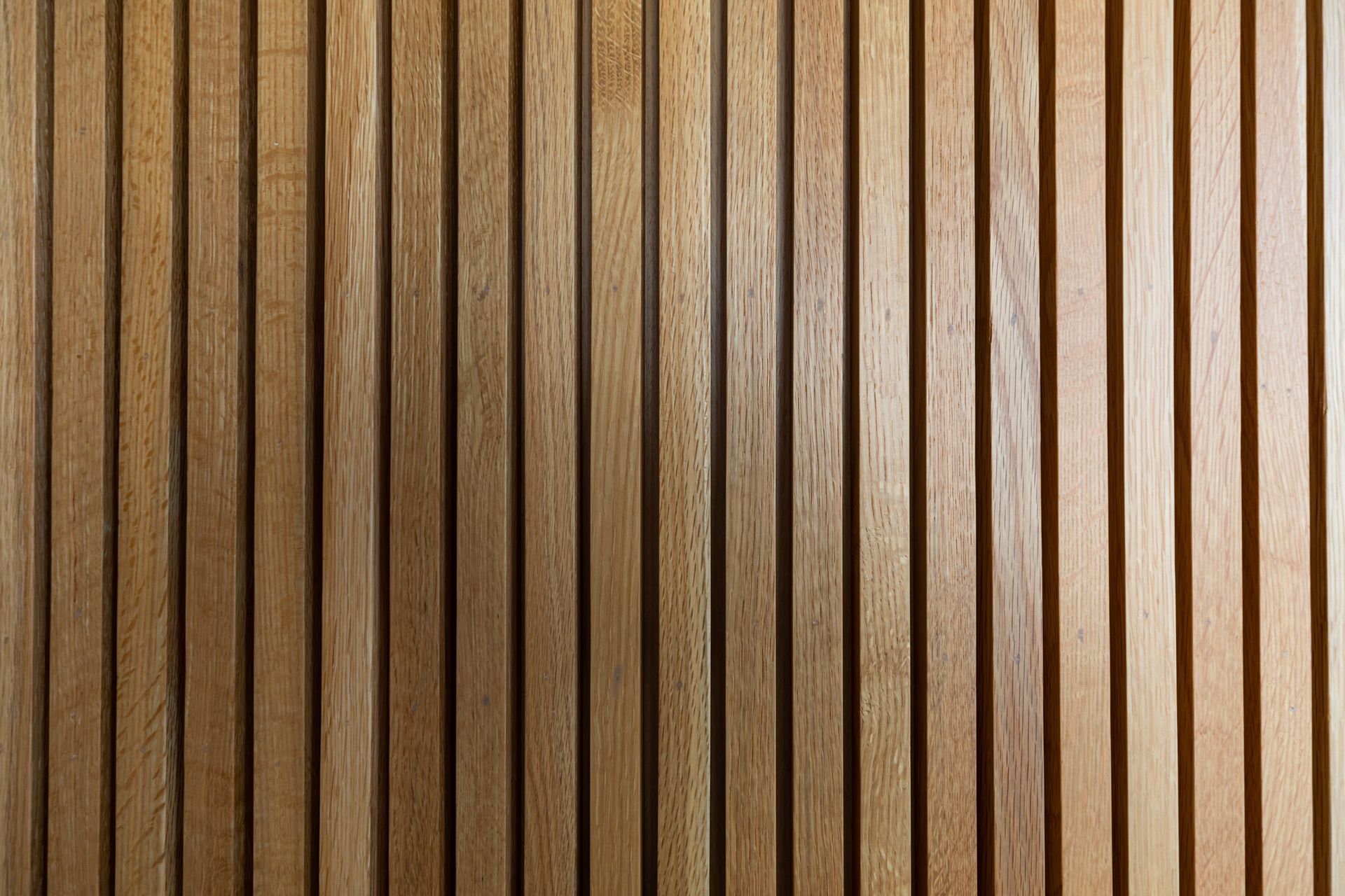 Gros plan sur un mur recouvert de lattes de bois verticales brun clair, entre lesquelles se projettent des ombres.