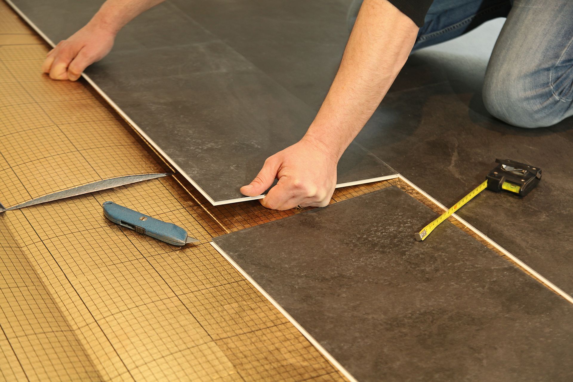 Une personne agenouillée sur le sol, en train d'installer des carreaux de sol rectangulaires gris foncé sur une sous-couche, avec des outils à proximité.