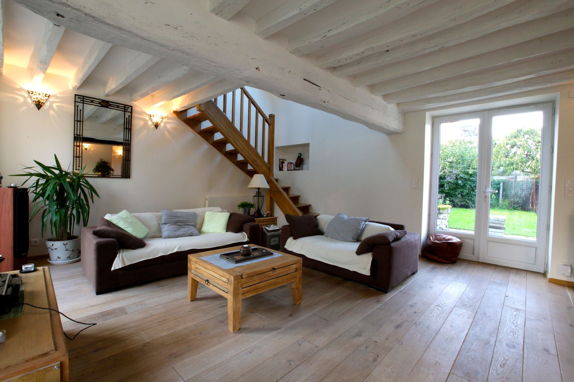 Salon avec parquet clair, plafond à poutres apparentes blanches, deux canapés marron, une table basse en bois et un escalier en bois.