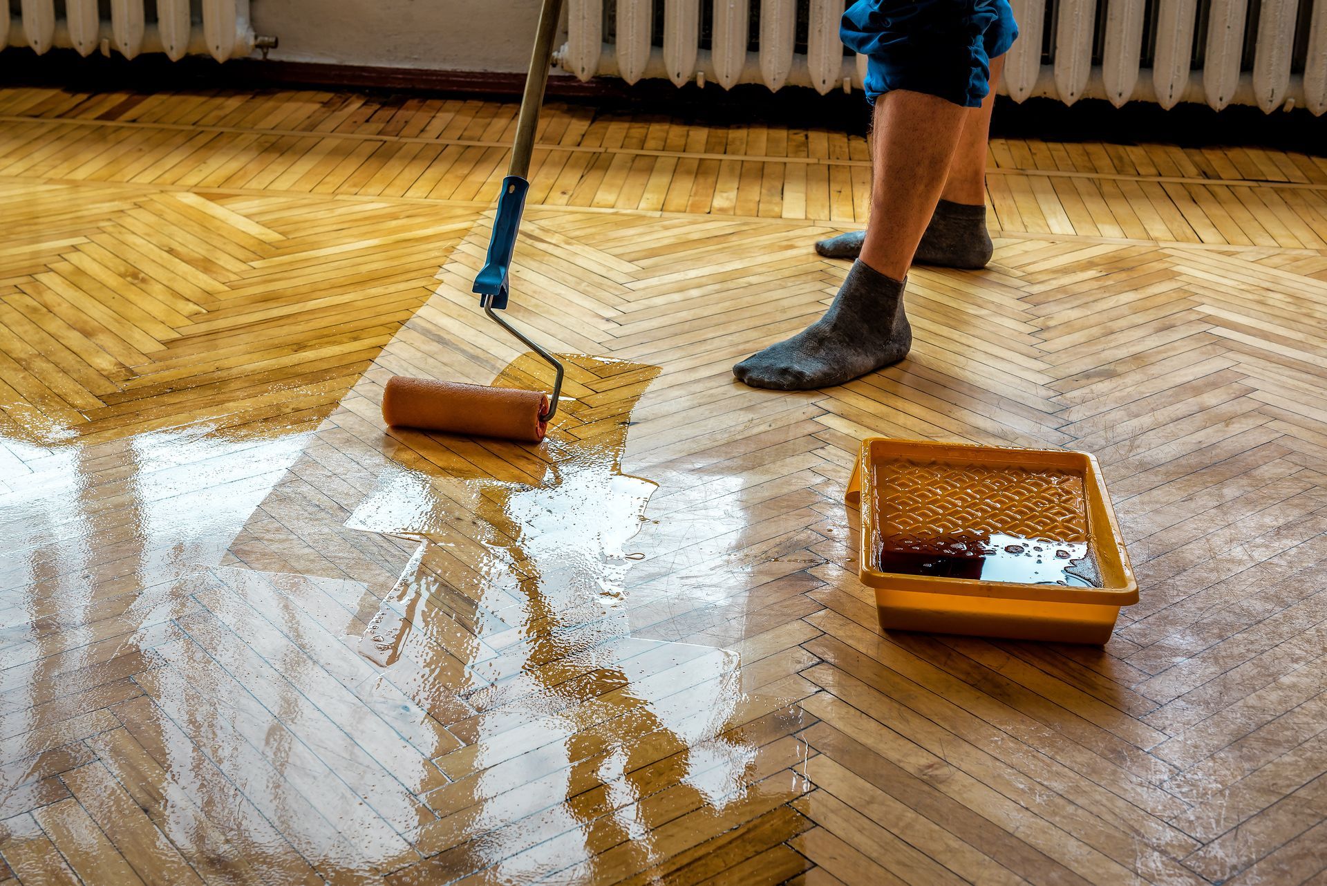 Une personne utilise un rouleau à peinture pour appliquer une finition transparente sur un parquet à chevrons, un bac à peinture étant posé à proximité.