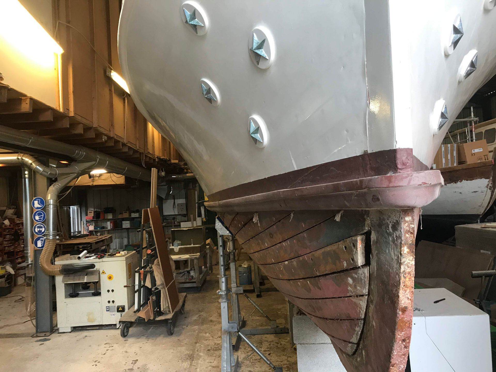 La proue d'un bateau en réparation, montrant une coque supérieure blanche ornée d'étoiles décoratives et une coque inférieure en bois foncé, dans un atelier.