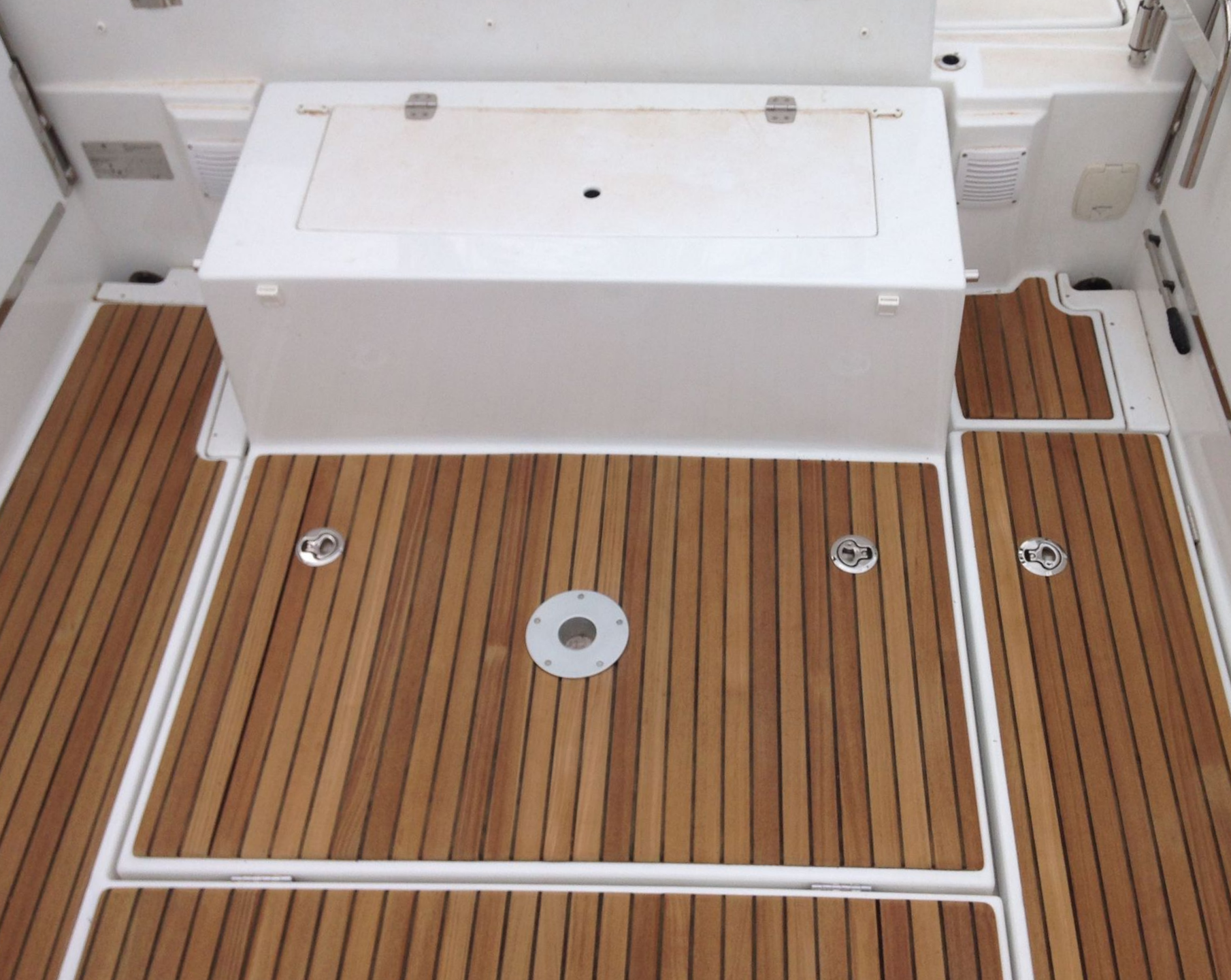Un pont en bois de teck sur un bateau, comportant une trappe de rangement rectangulaire centrale, un support sur piédestal et un grand coffre de rangement blanc.