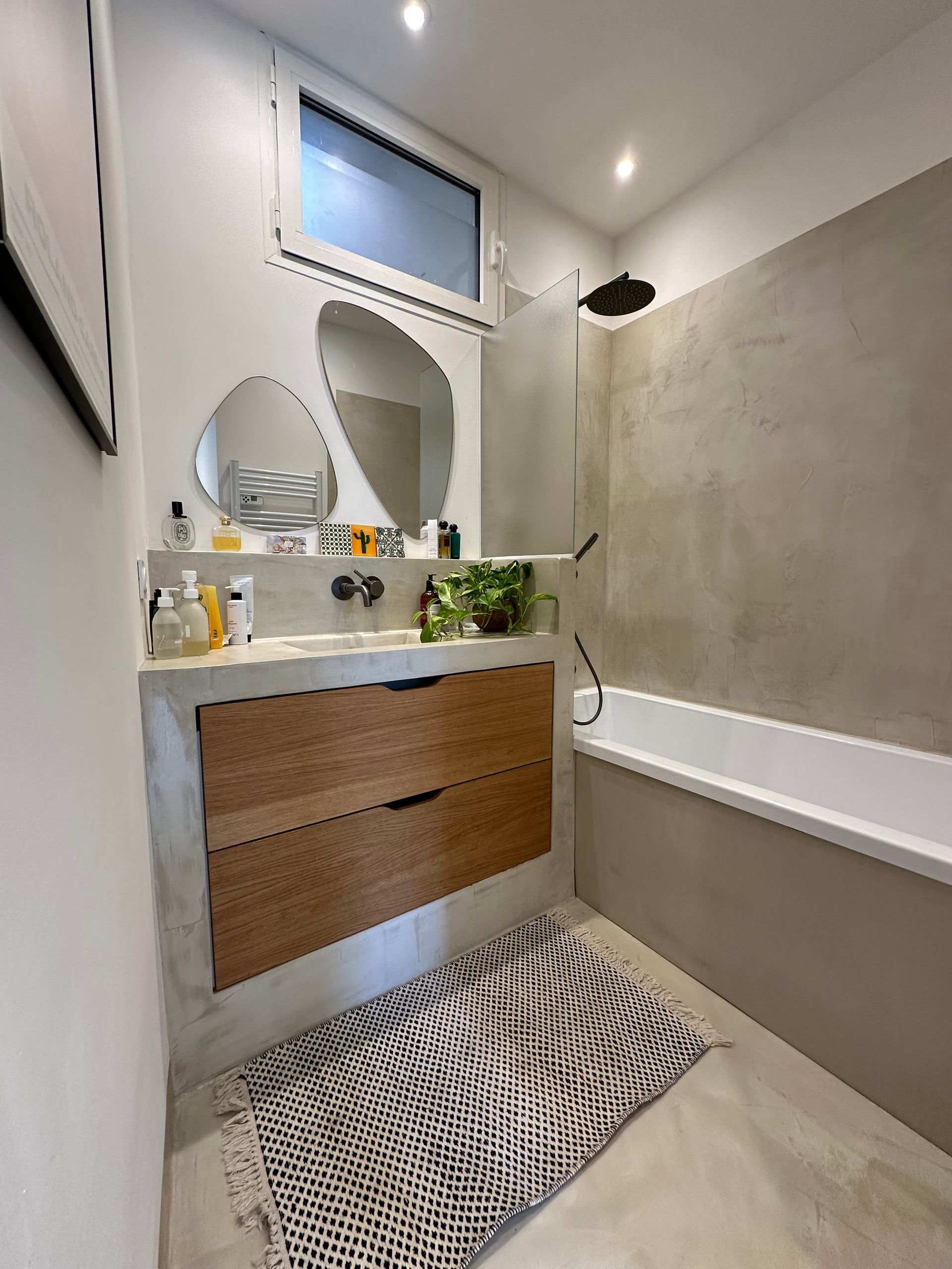Une salle de bains moderne avec un meuble-lavabo en bois, deux miroirs de forme irrégulière, une baignoire-douche et un tapis à motifs.