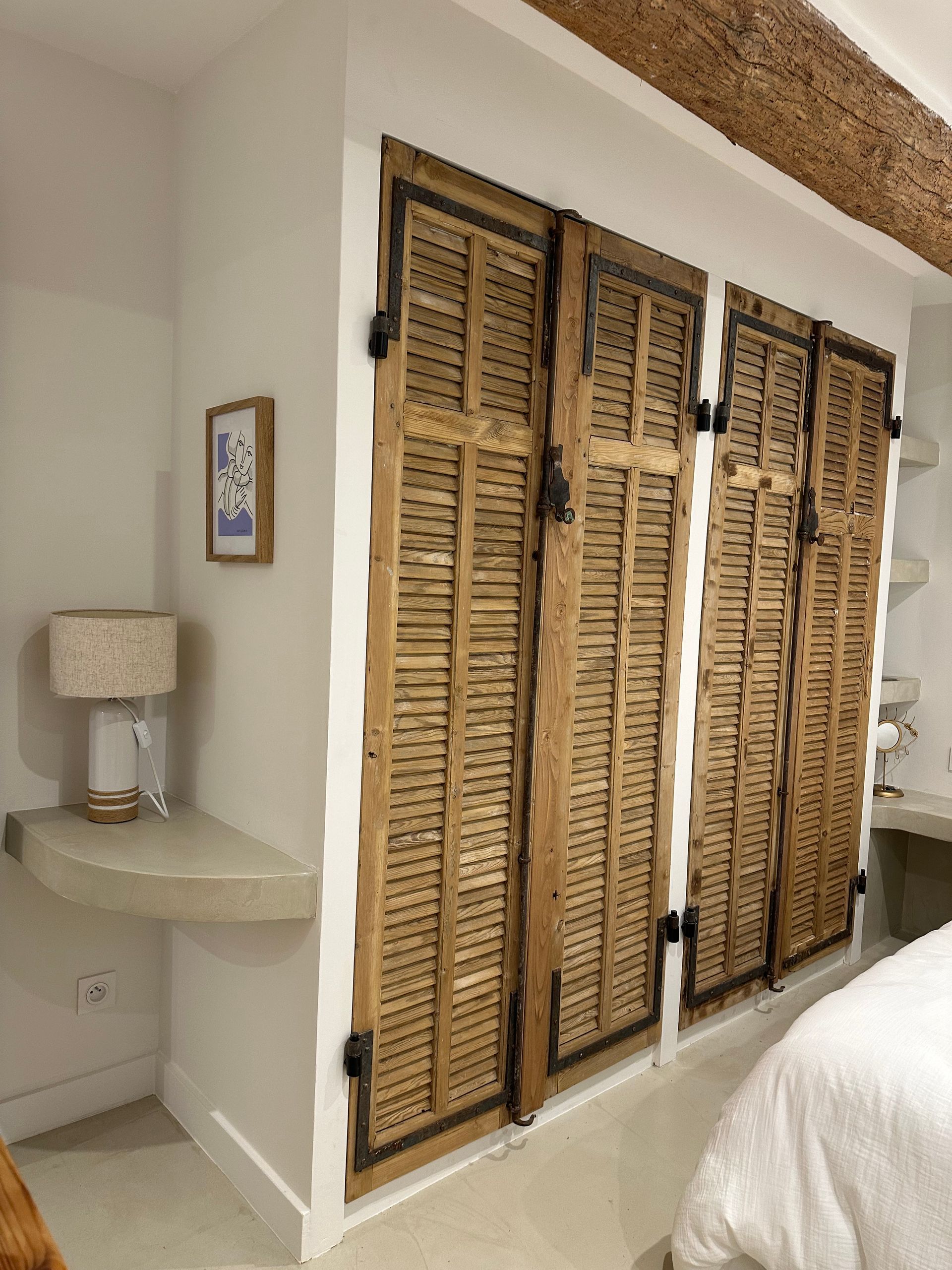 Un coin de chambre avec une étagère en pierre flottante sur laquelle repose une lampe, à côté d'un mur orné de volets rustiques en bois.