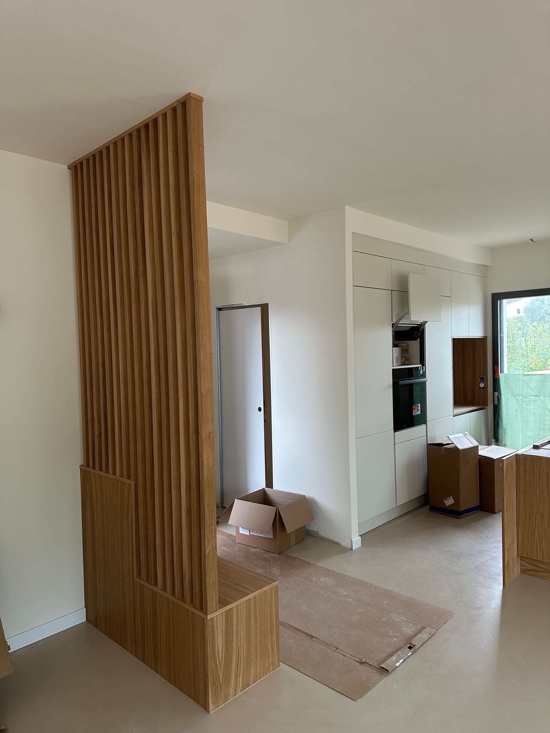 Une cloison en lattes de bois verticales sépare une cuisine partiellement aménagée, avec des meubles et des boîtes blancs.