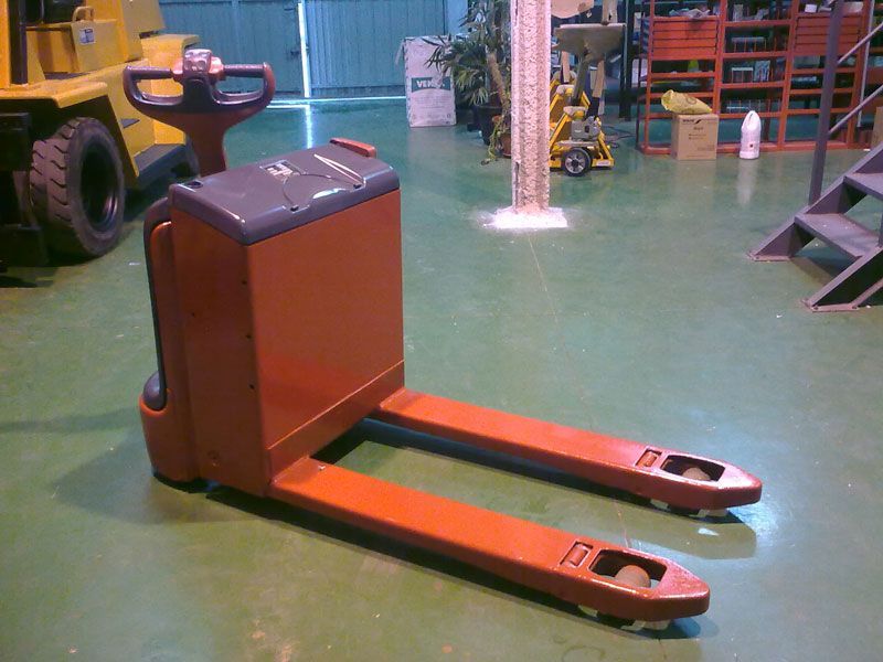 Una transpaleta naranja está sobre un suelo verde en un almacén.