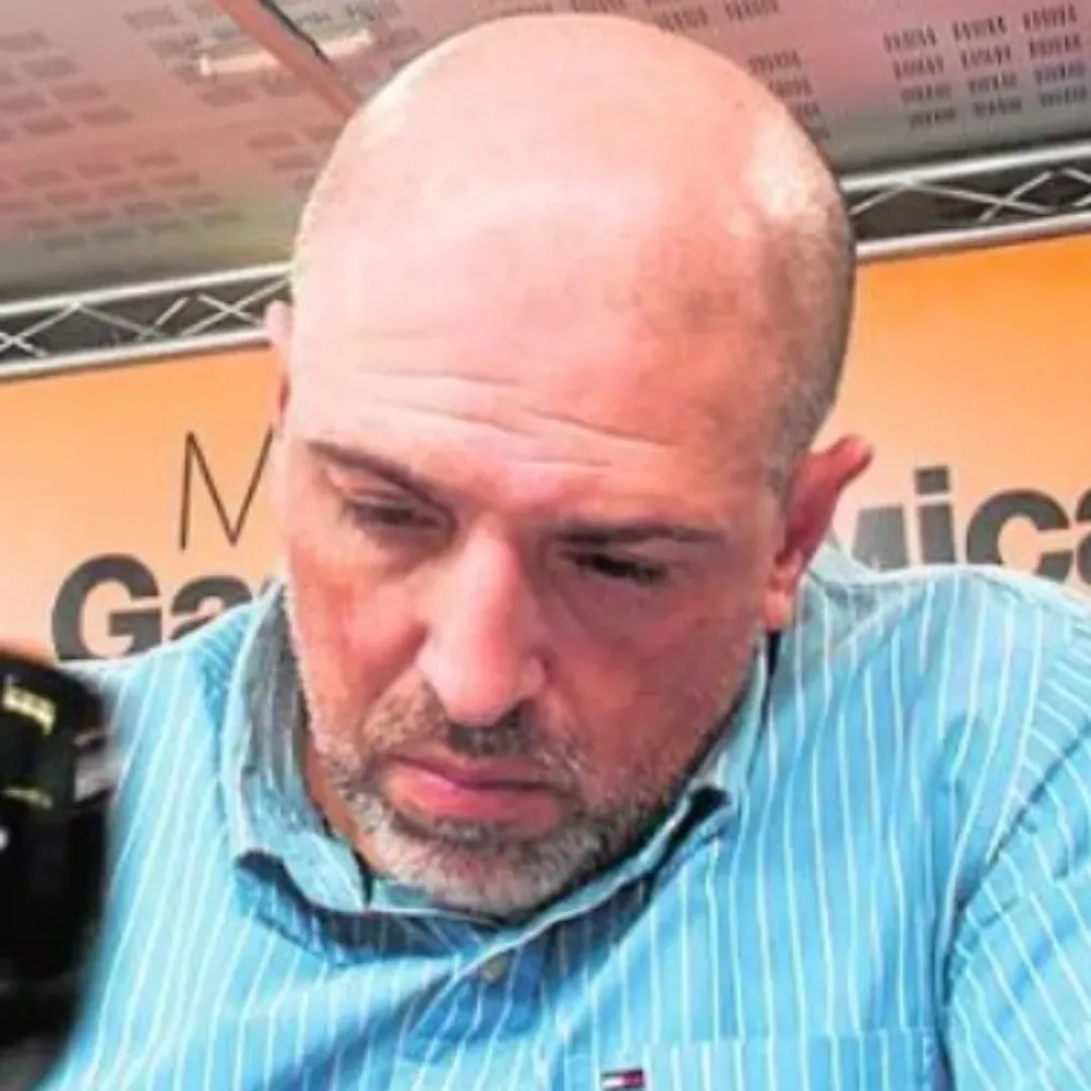 Un hombre calvo con barba corta, que viste una camisa abotonada de rayas azul claro, mira hacia abajo.