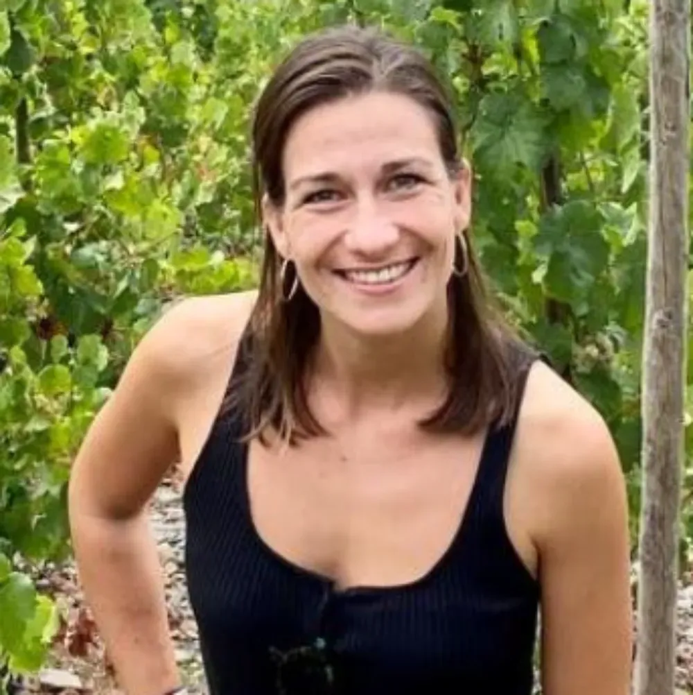 Una persona sonriente, vestida con una camiseta sin mangas negra y pendientes de aro, de pie frente a un viñedo frondoso.