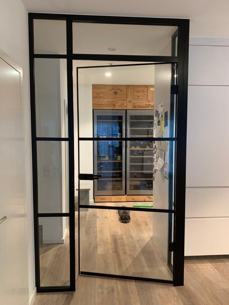 Eine schwarz umrahmte Glastür ist geöffnet und gibt den Blick auf eine Küche mit Holzschränken und -geräten frei.