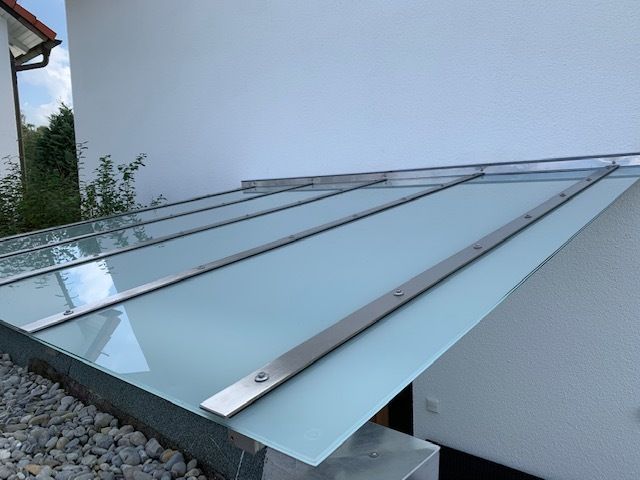 Eine Markise aus Glas und Metall über einem Außeneingang, der an ein weißes Gebäude angebaut ist.