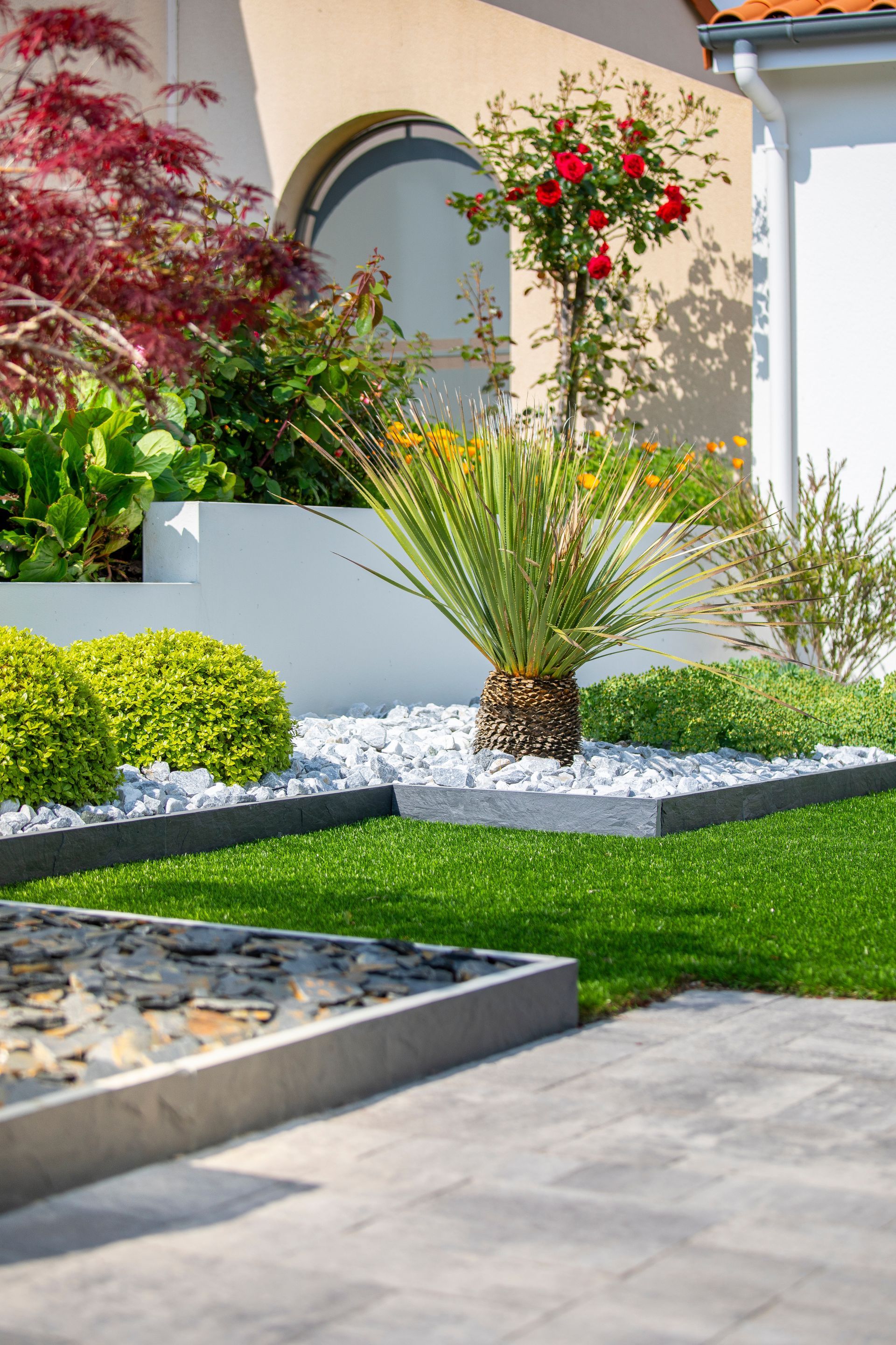 Jardin bien entretenu avec de l'herbe verte, des rochers blancs et diverses plantes devant une maison.