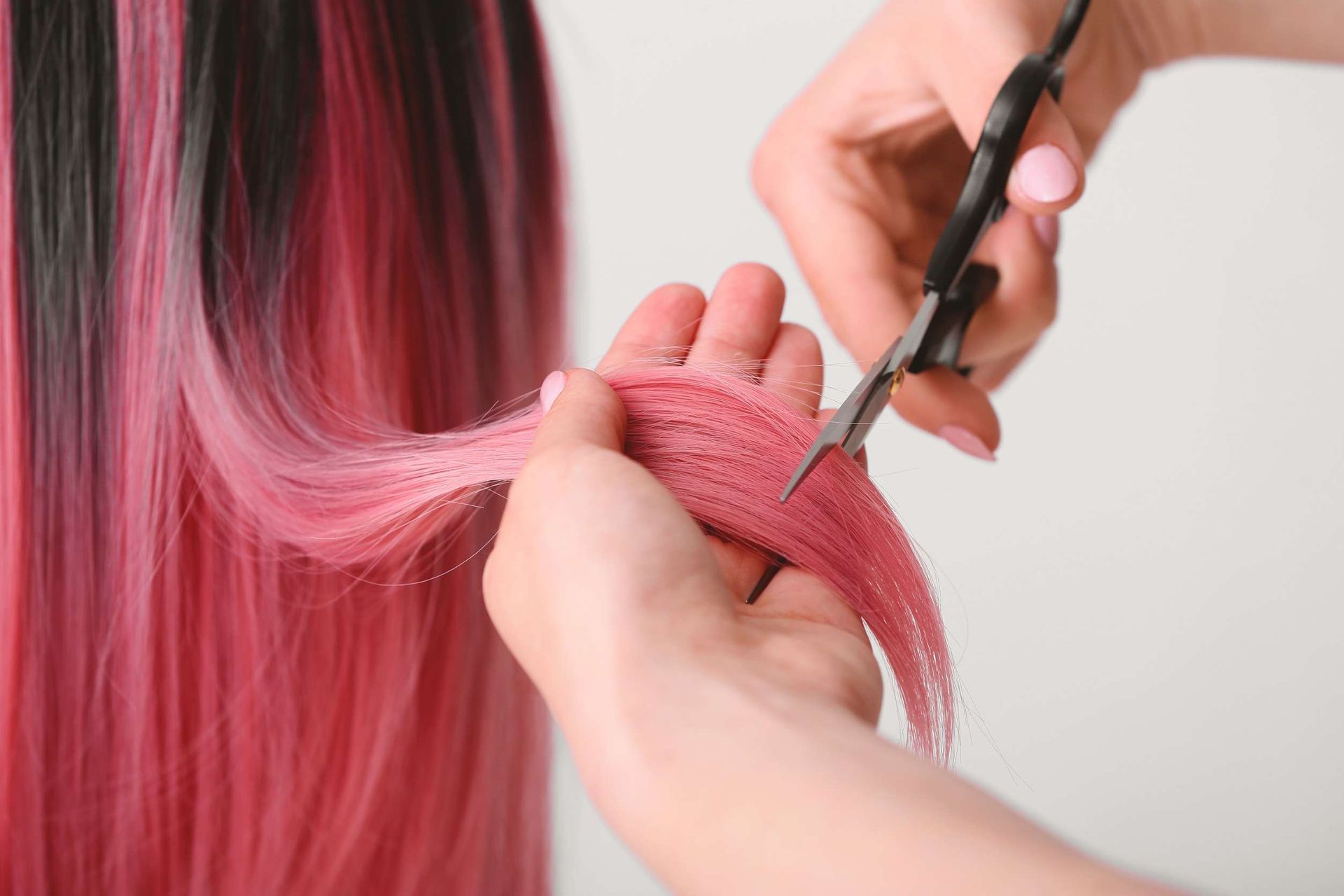 Manos cortando cabello rosa con tijeras negras.